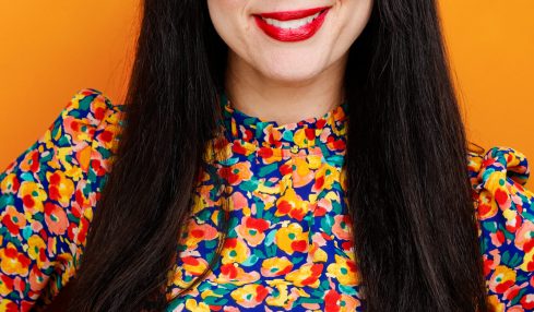 Woman with long dark hair, red lipstick, smiling, wearing a colorful patterned top, standing against an orange background.
