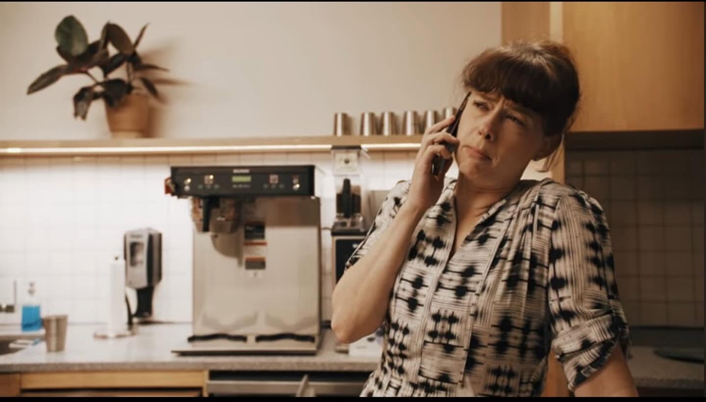 Person with short dark hair talking on a phone in a kitchen, wearing a patterned shirt, with kitchen appliances behind.