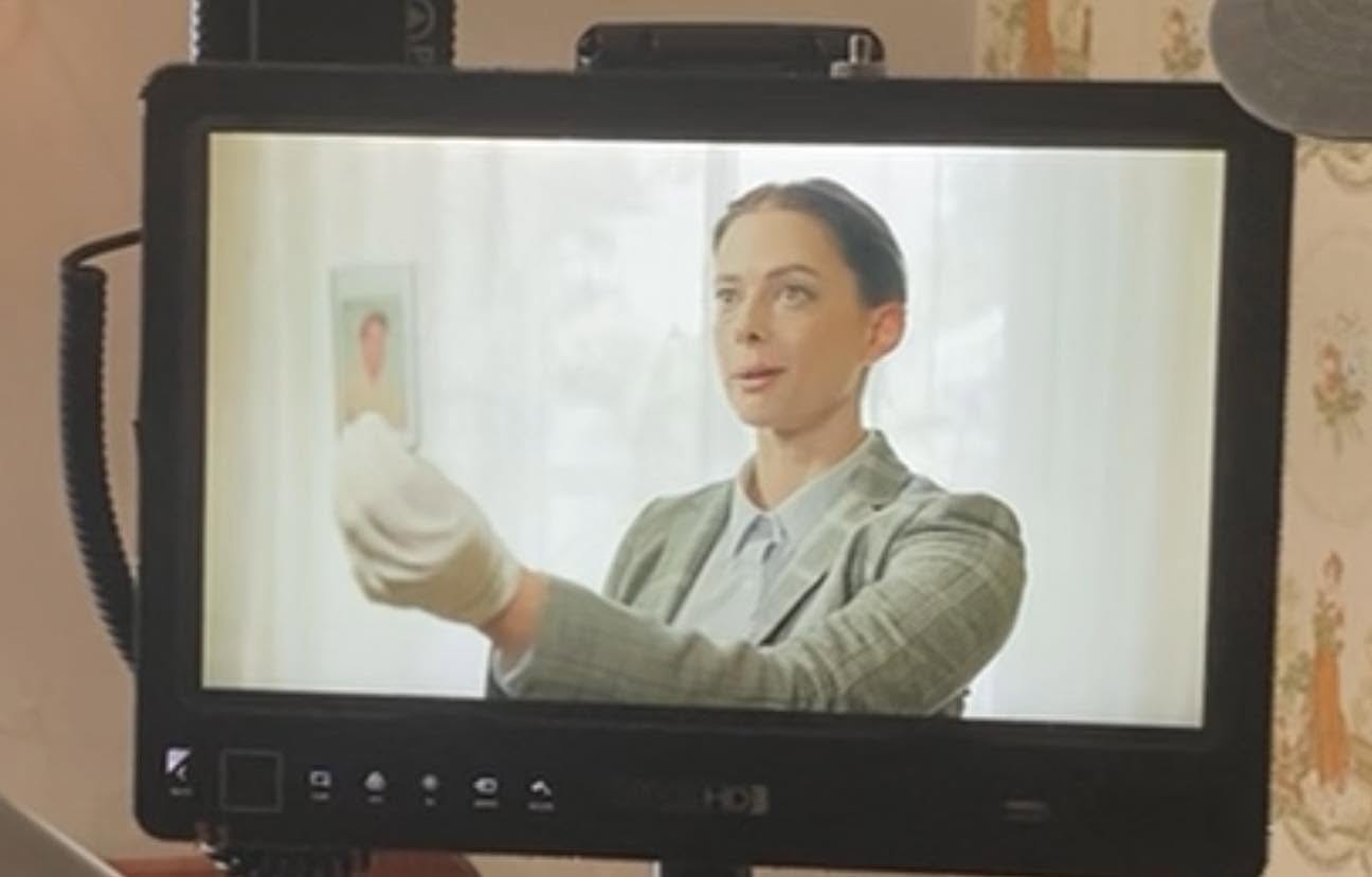 A woman in a gray blazer holds a small object in her gloved hand, standing in a bright room with a window.