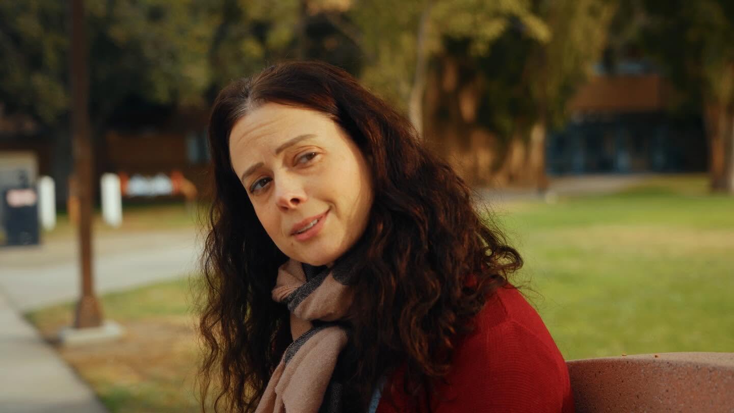 Woman with dark curly hair sitting outdoors in a park, wearing a red top and a scarf, smiling slightly.