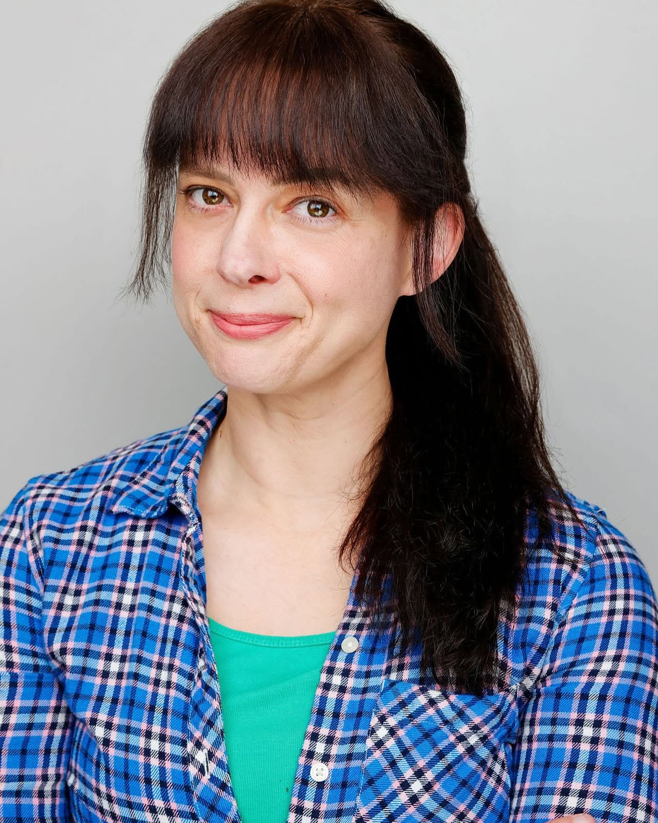 Woman with dark brown hair and bangs, wearing a blue plaid shirt over a green top, smiling softly.