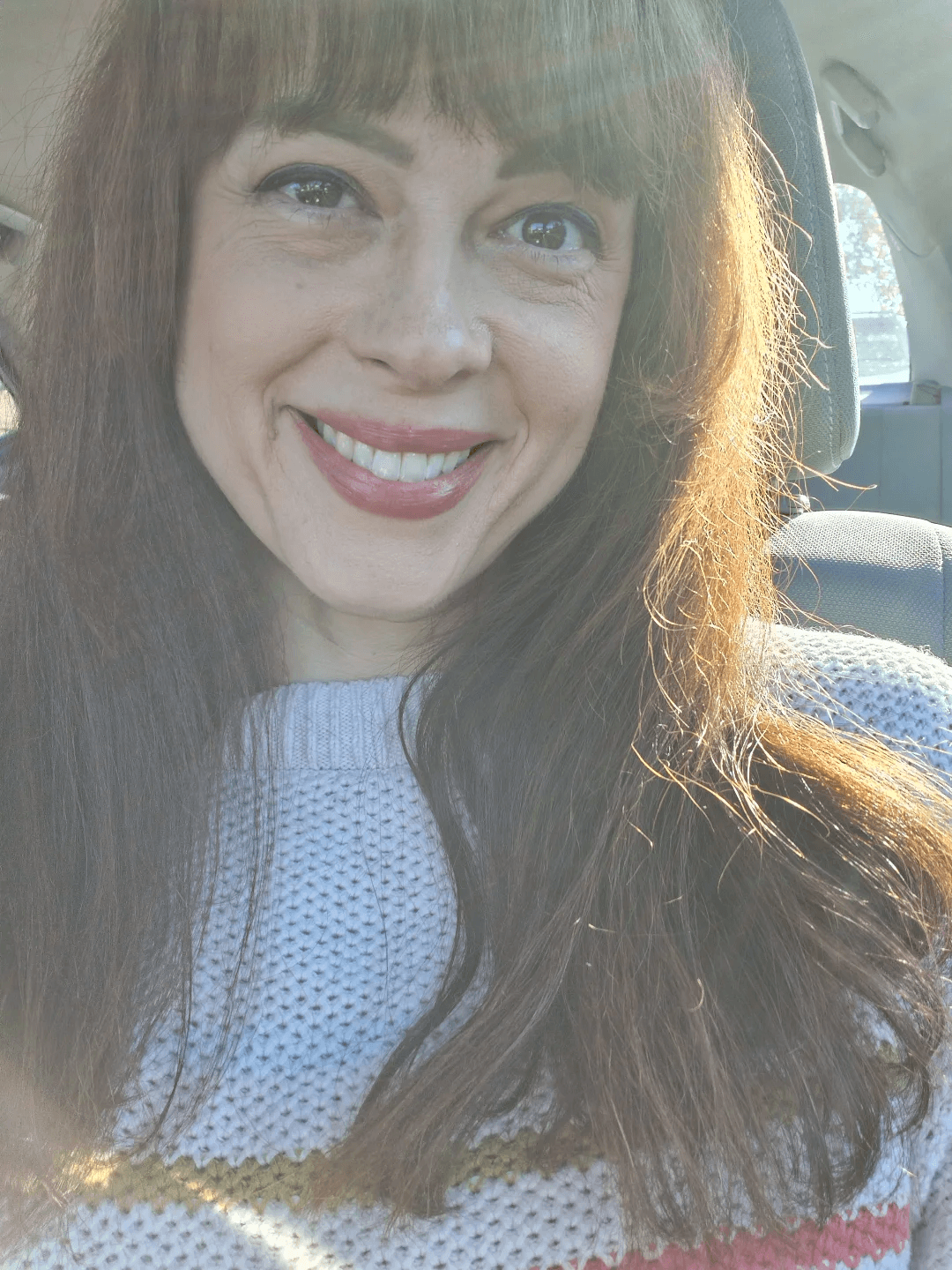 Smiling woman with long brown hair and bangs, wearing a light-colored sweater, sitting in a car with sunlight on her face.