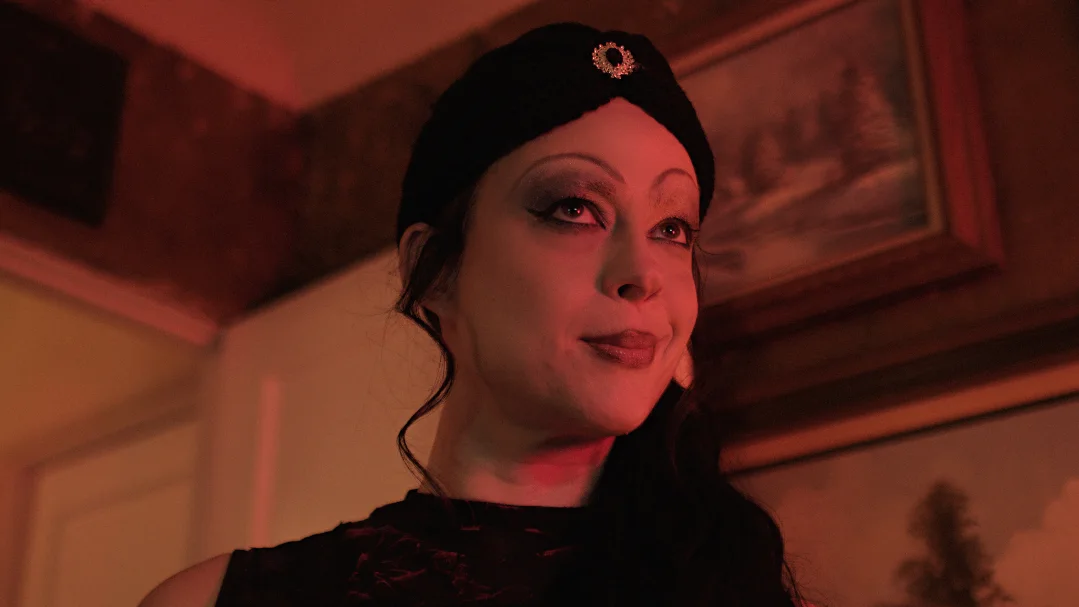 Woman with dark hair and makeup, wearing a black headband with a decorative element, in a warmly lit room with framed artwork.