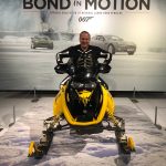 Person sitting on a yellow snowmobile in front of a 'Bond in Motion' display with cars and smoke in the background.
