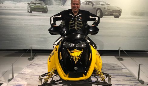Person sitting on a yellow snowmobile in front of a 'Bond in Motion' display with cars and smoke in the background.