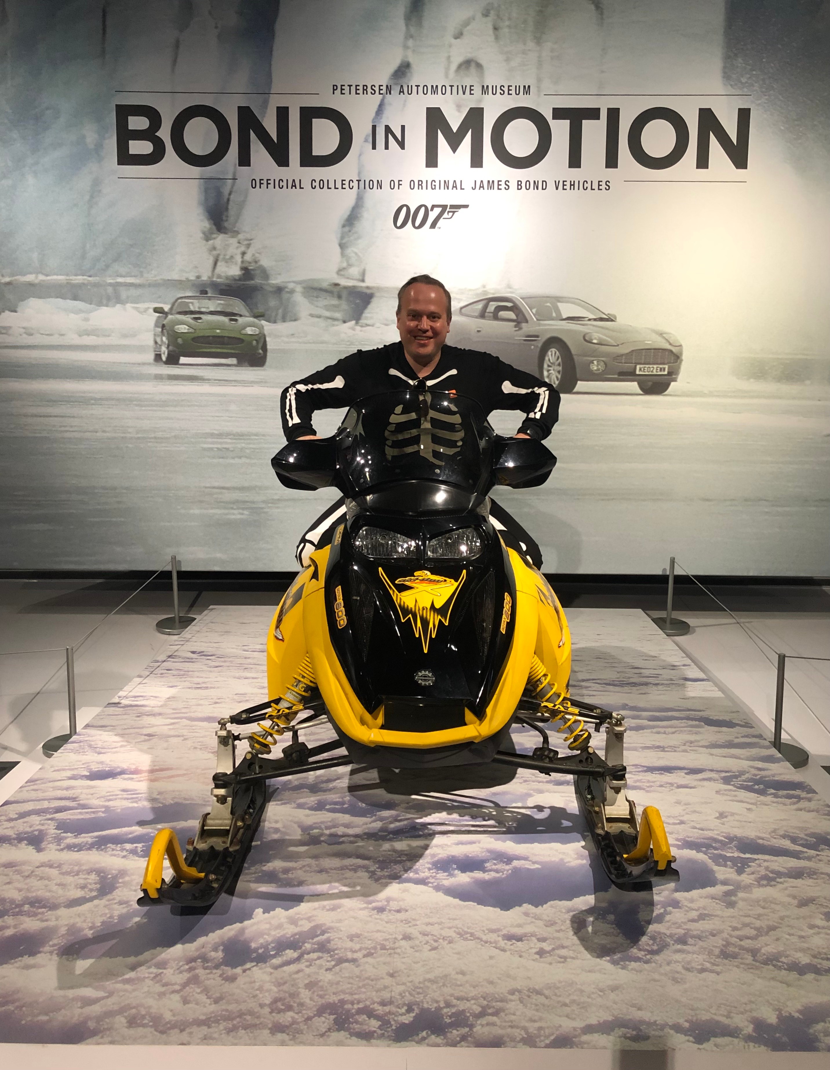 Person sitting on a yellow snowmobile in front of a 'Bond in Motion' display with cars and smoke in the background.