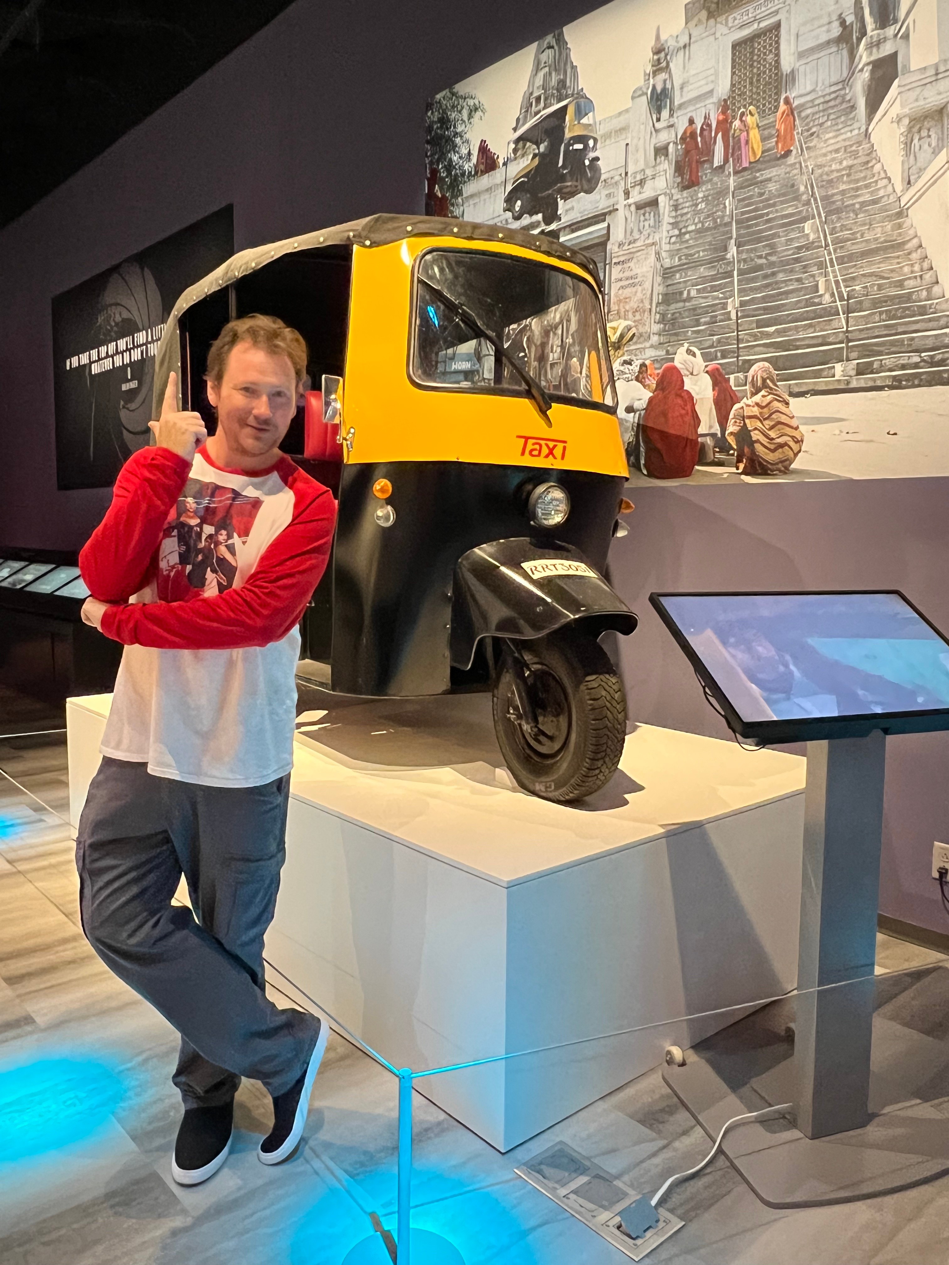 Person standing next to a yellow and black auto rickshaw display at a museum, with a large screen nearby.