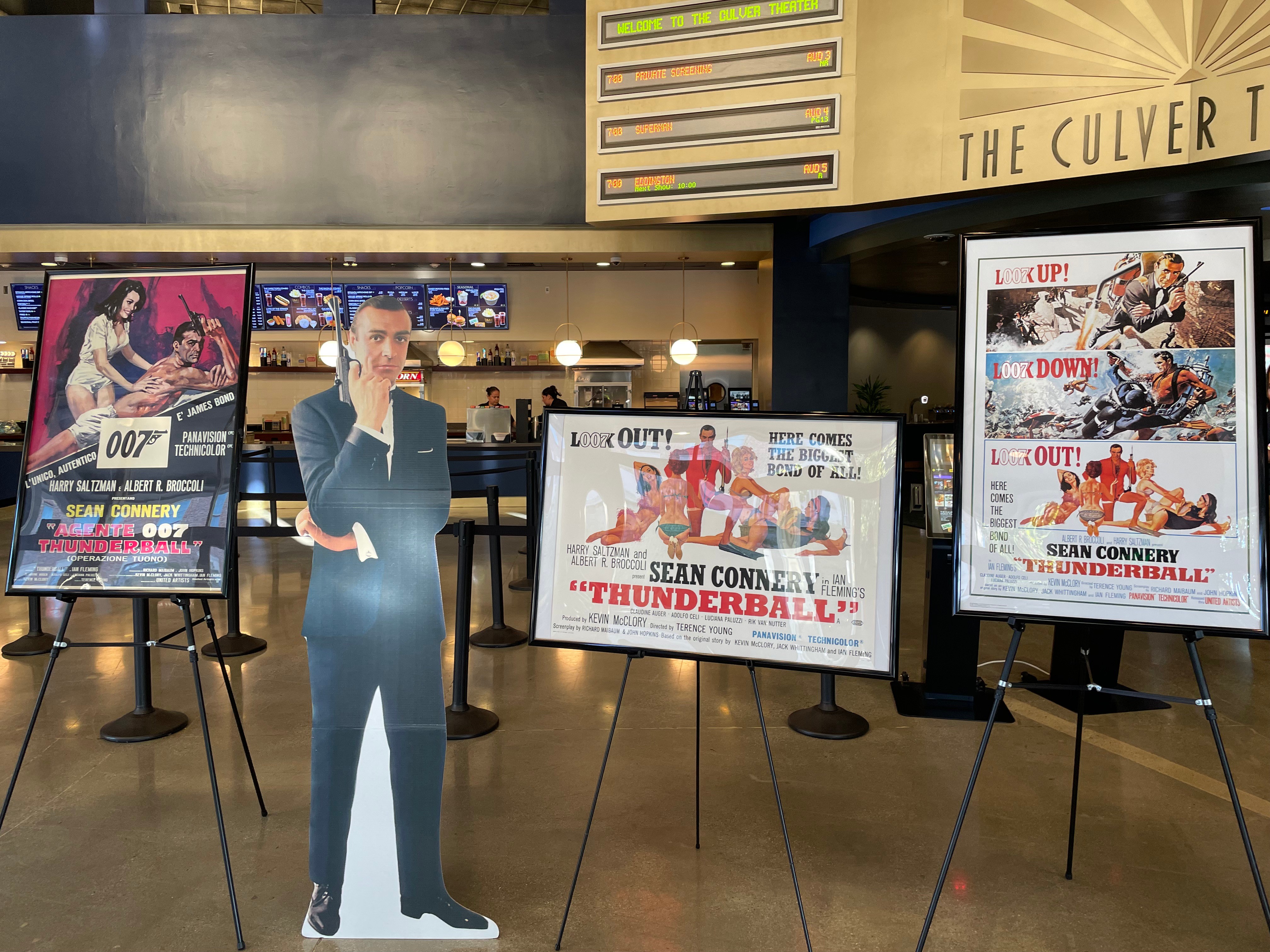 Four movie posters and a life-sized cutout of a man in a suit inside a theater lobby.
