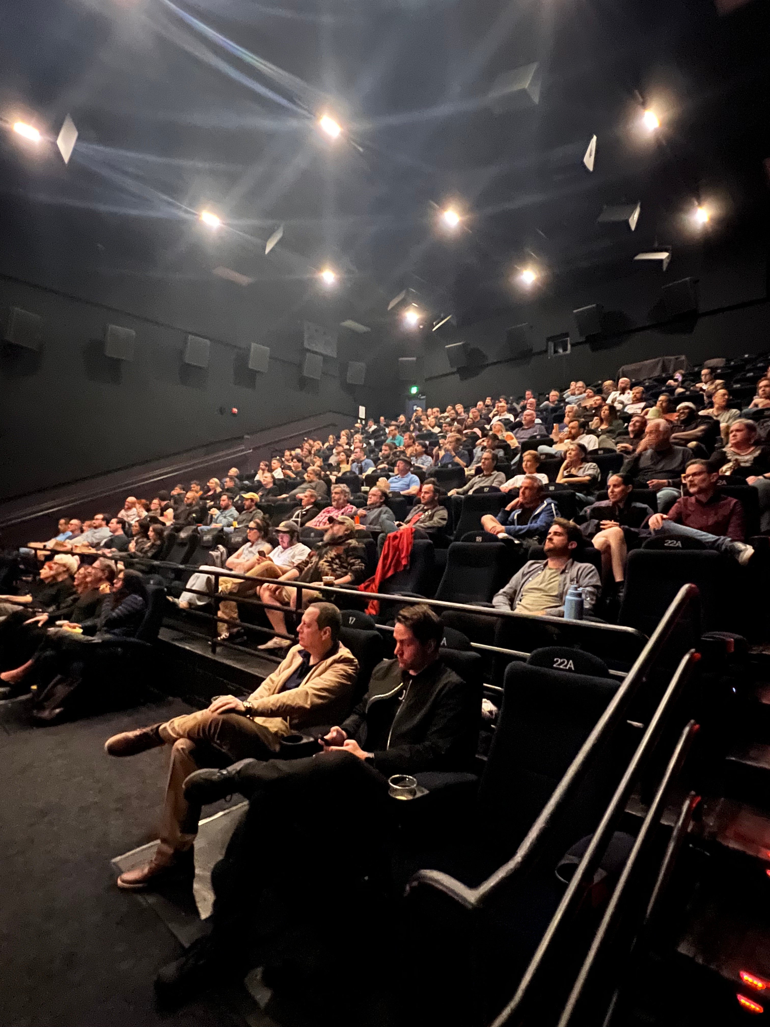 Audience seated in a dark theater, facing a stage with bright overhead lights, some people watching attentively.