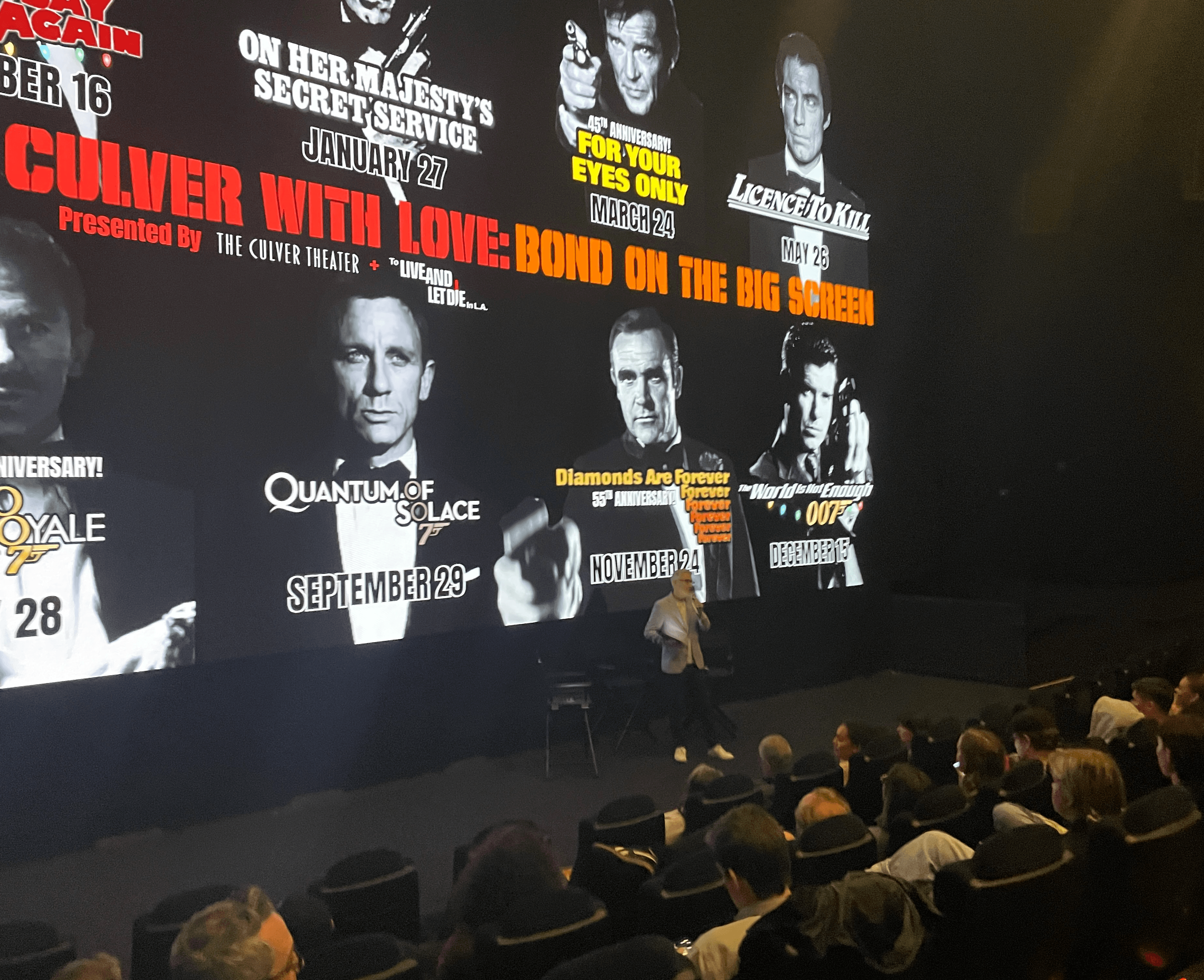 Movie theater screen displaying promotional images and text for a film event, with audience seated in front.