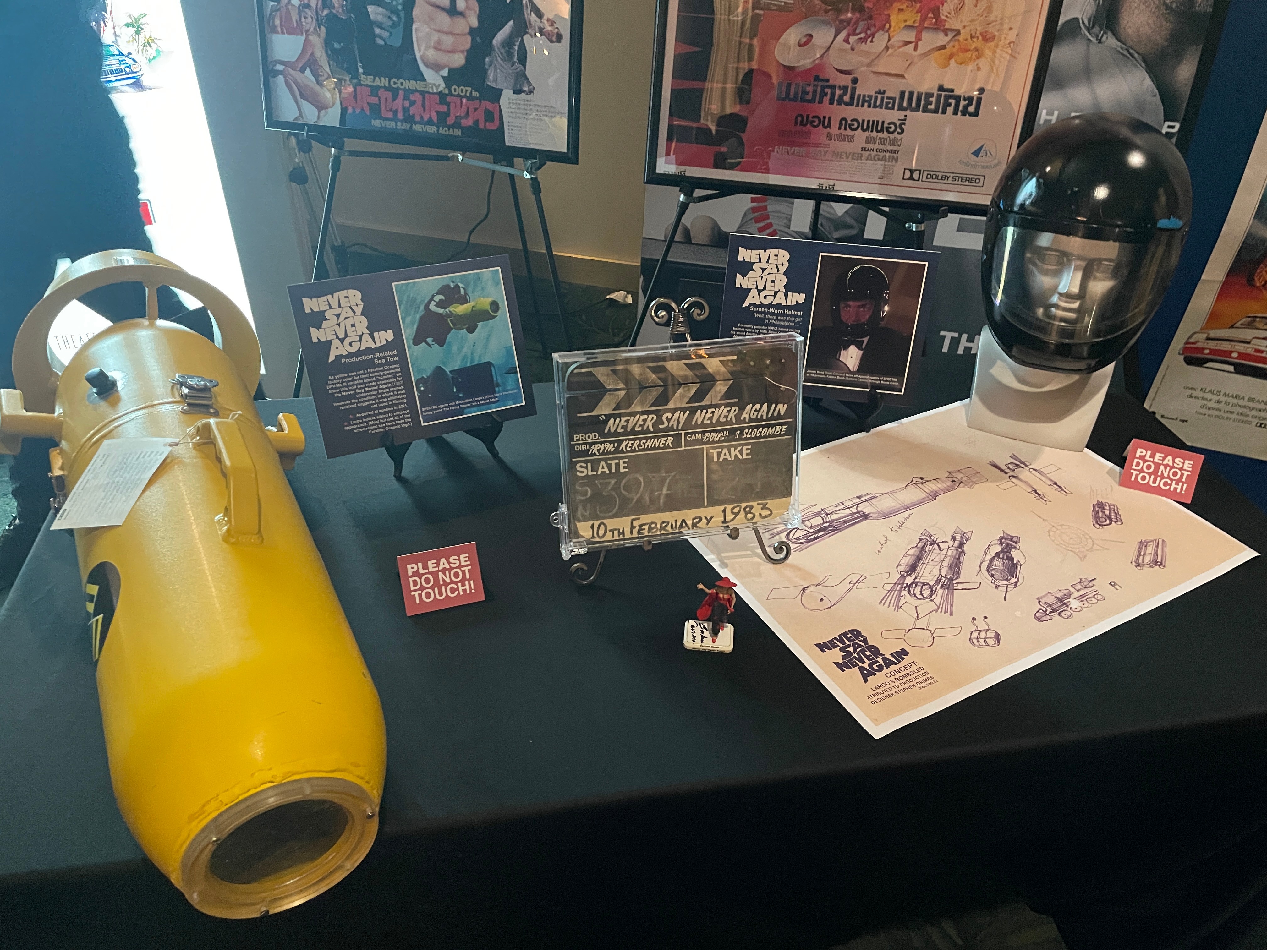 Display table with movie memorabilia, sketches, and a helmet, with screens and posters in the background.
