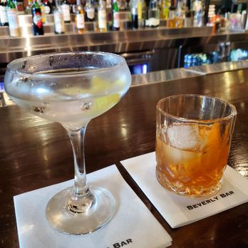 Two cocktails on a bar counter, one in a coupe glass with a lemon slice, the other in a rocks glass with an orange peel, Beverly Bar napkins, bar bottles in background.