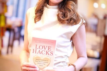 Woman smiling holding a book titled 'Love Hacks' in a cozy indoor setting.