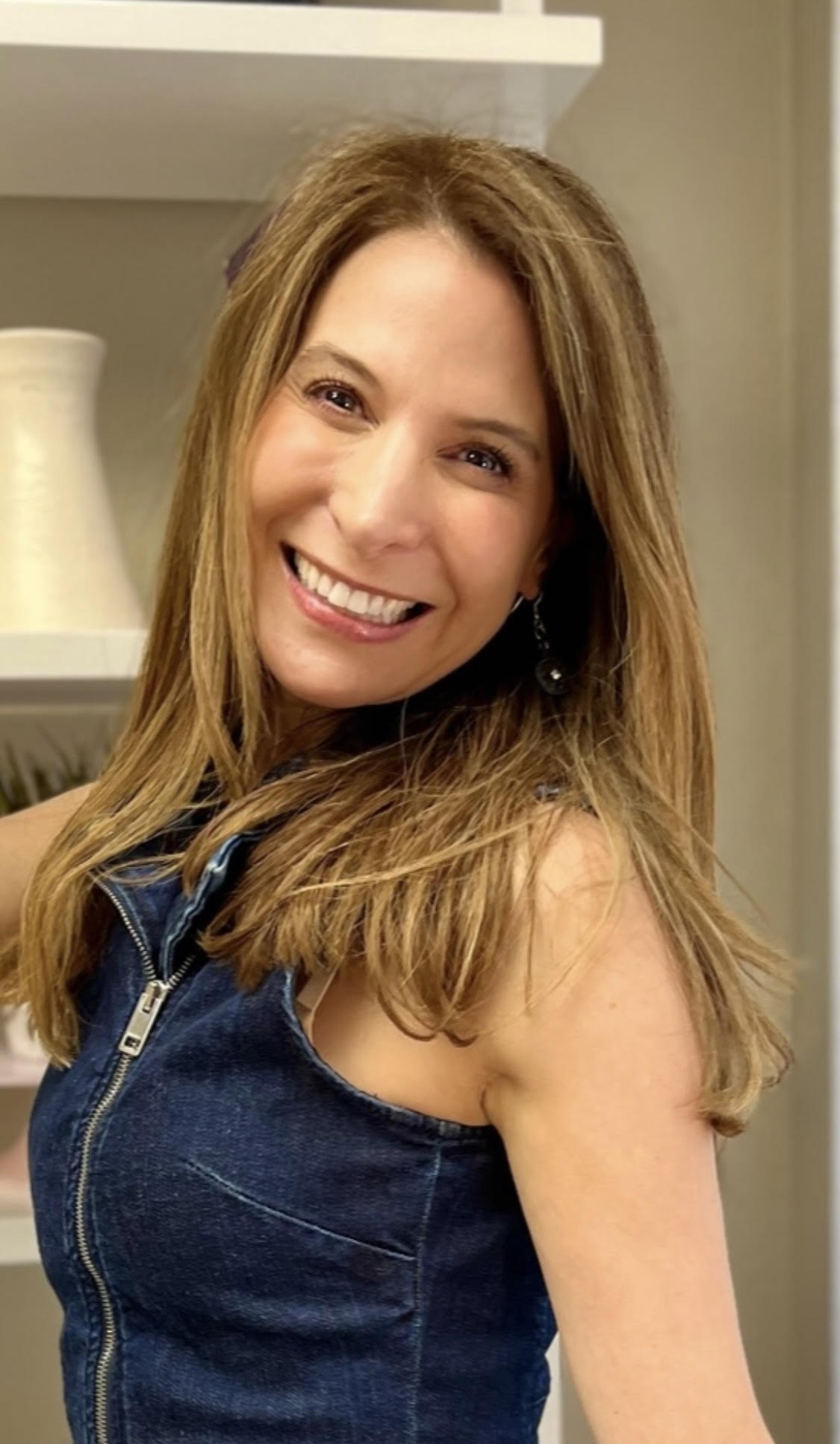 Smiling woman with long hair wearing a sleeveless denim top, standing indoors near a shelf with a vase.