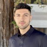 Young man with short dark hair and light skin, wearing a black collared shirt, standing outdoors near a tree.