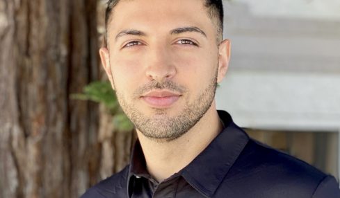 Young man with short dark hair and light skin, wearing a black collared shirt, standing outdoors near a tree.
