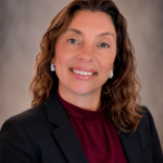 Portrait of a woman with wavy brown hair, smiling, wearing a black blazer and maroon blouse, against a neutral background.