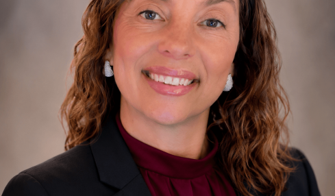 Portrait of a woman with wavy brown hair, smiling, wearing a black blazer and maroon blouse, against a neutral background.