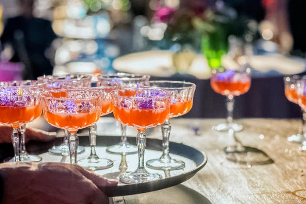 Tray of pink and purple cocktails on a table with blurred background, possibly at a party or event.