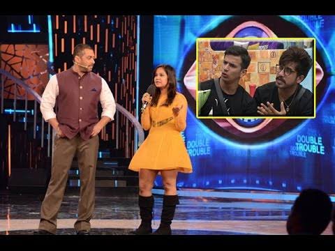 Three people on a stage, woman in yellow dress speaking, two men listening, inset shows two young men, colorful background