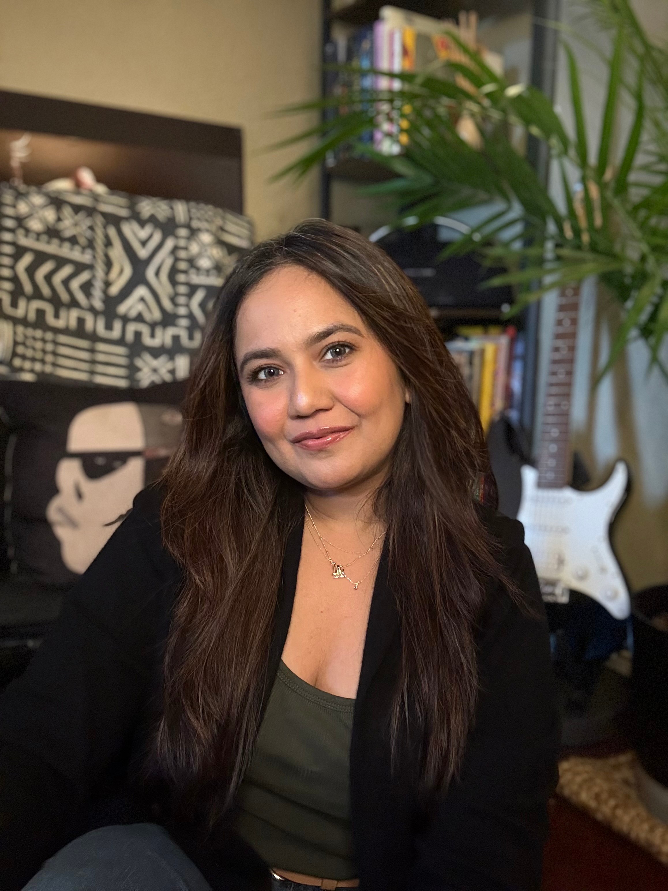 Woman with long dark hair smiling indoors, background includes a bed, bookshelf, and guitar.