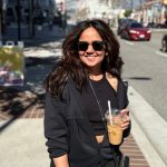 Woman with sunglasses smiling holding a drink on a city sidewalk, sunny day, buildings and cars in background.