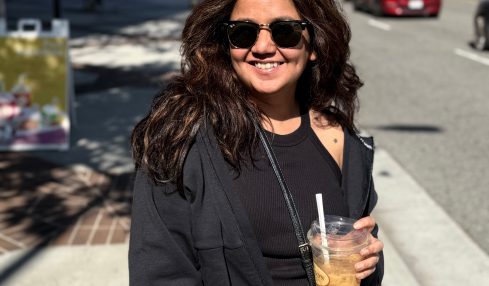 Woman with sunglasses smiling holding a drink on a city sidewalk, sunny day, buildings and cars in background.
