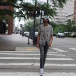 Person walking across a city crosswalk with trees and buildings in the background, wearing a patterned shirt, jeans, and sneakers.