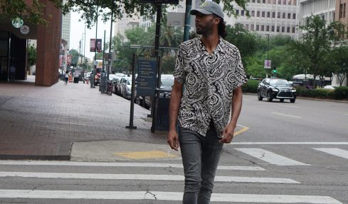 Person walking across a city crosswalk with trees and buildings in the background, wearing a patterned shirt, jeans, and sneakers.