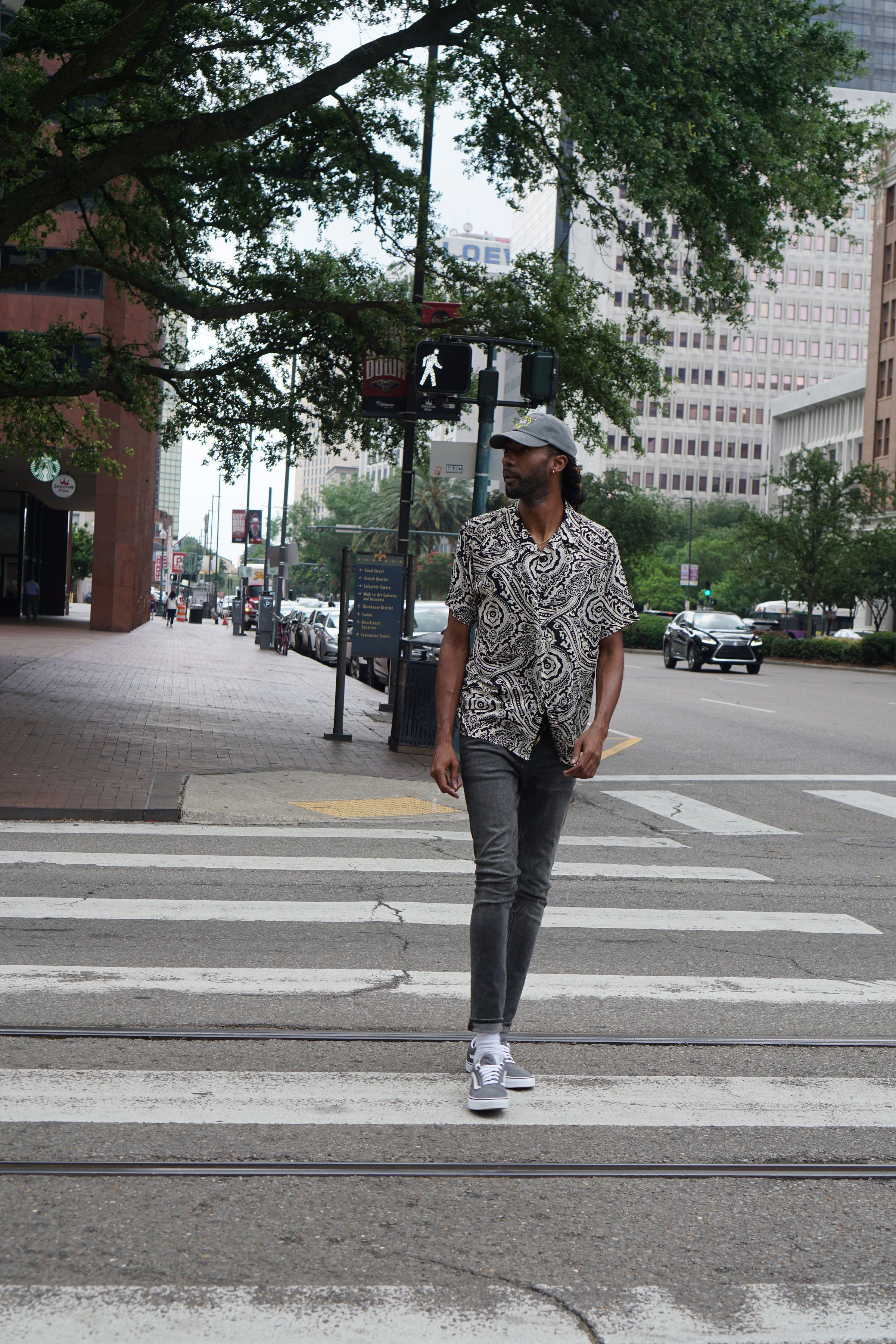 Person walking across a city crosswalk with trees and buildings in the background, wearing a patterned shirt, jeans, and sneakers.