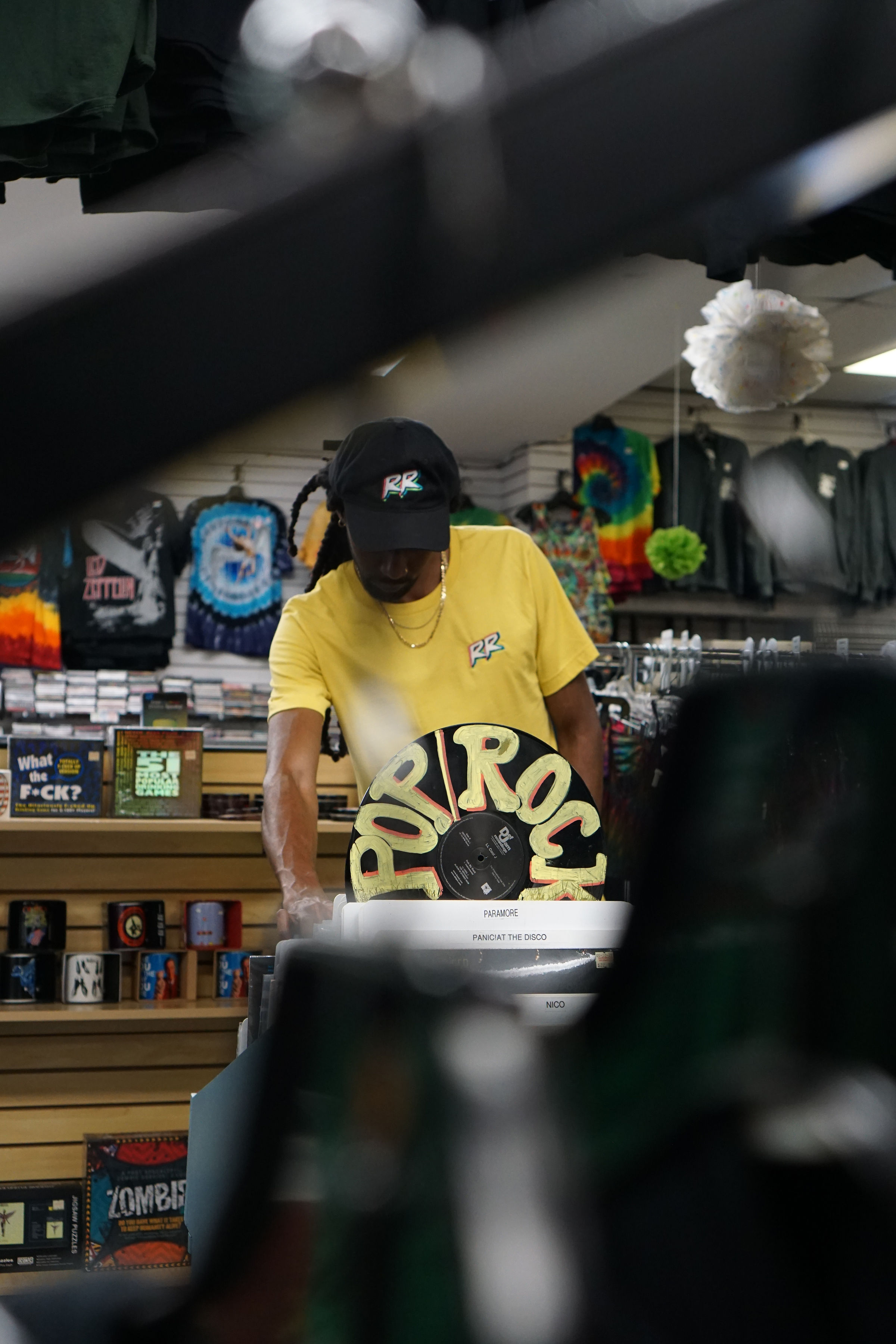 Person wearing a yellow shirt and black cap shopping in a store with colorful merchandise and decorations behind them.