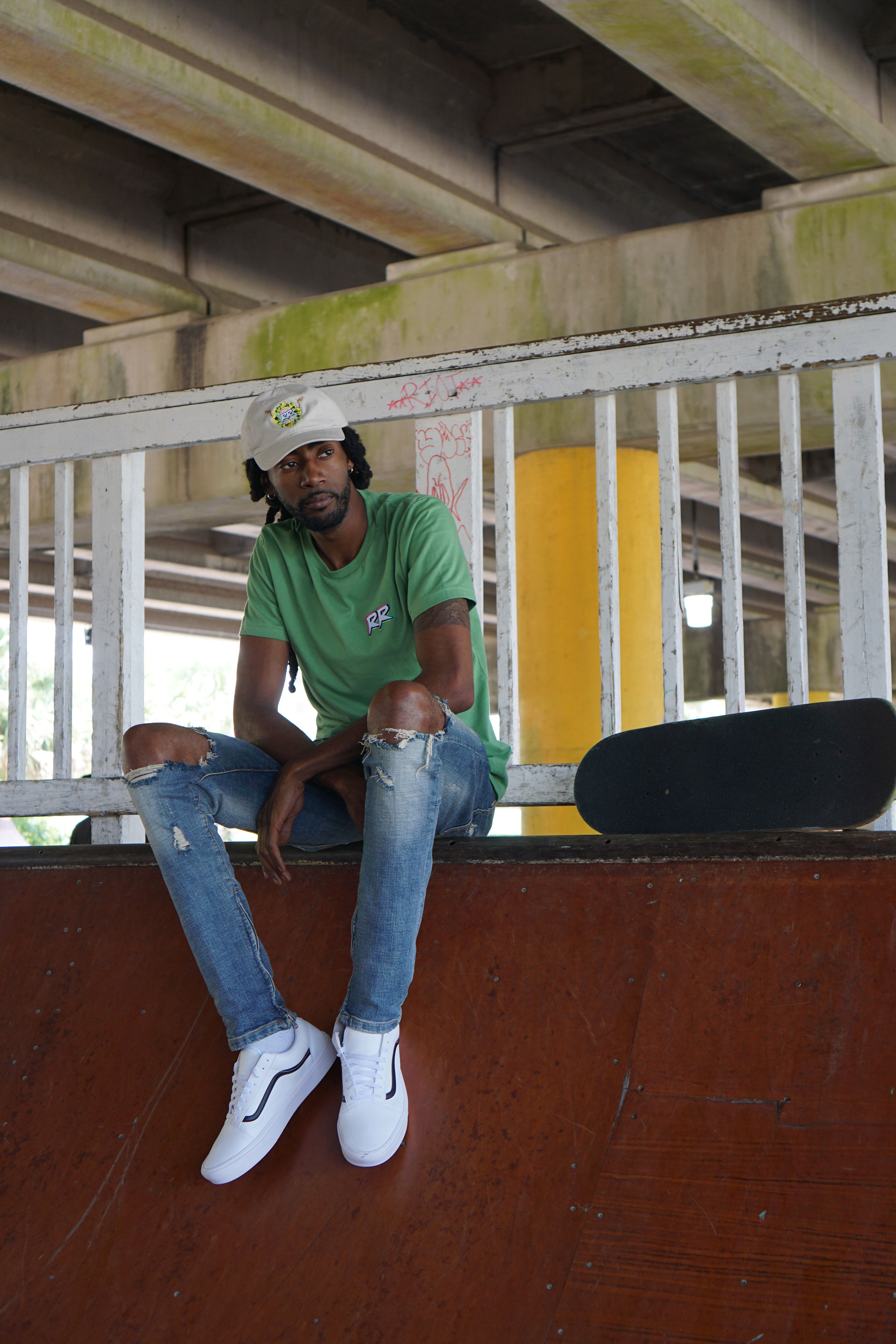 Young man sitting on a ledge under a bridge, wearing a green shirt, ripped jeans, white sneakers, and a cap.