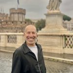 Man smiling outdoors near a fountain with a statue of an angel holding a cross, cloudy sky background.