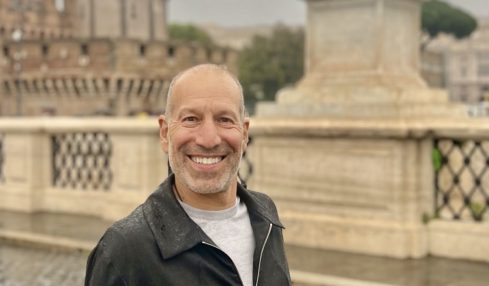 Man smiling outdoors near a fountain with a statue of an angel holding a cross, cloudy sky background.