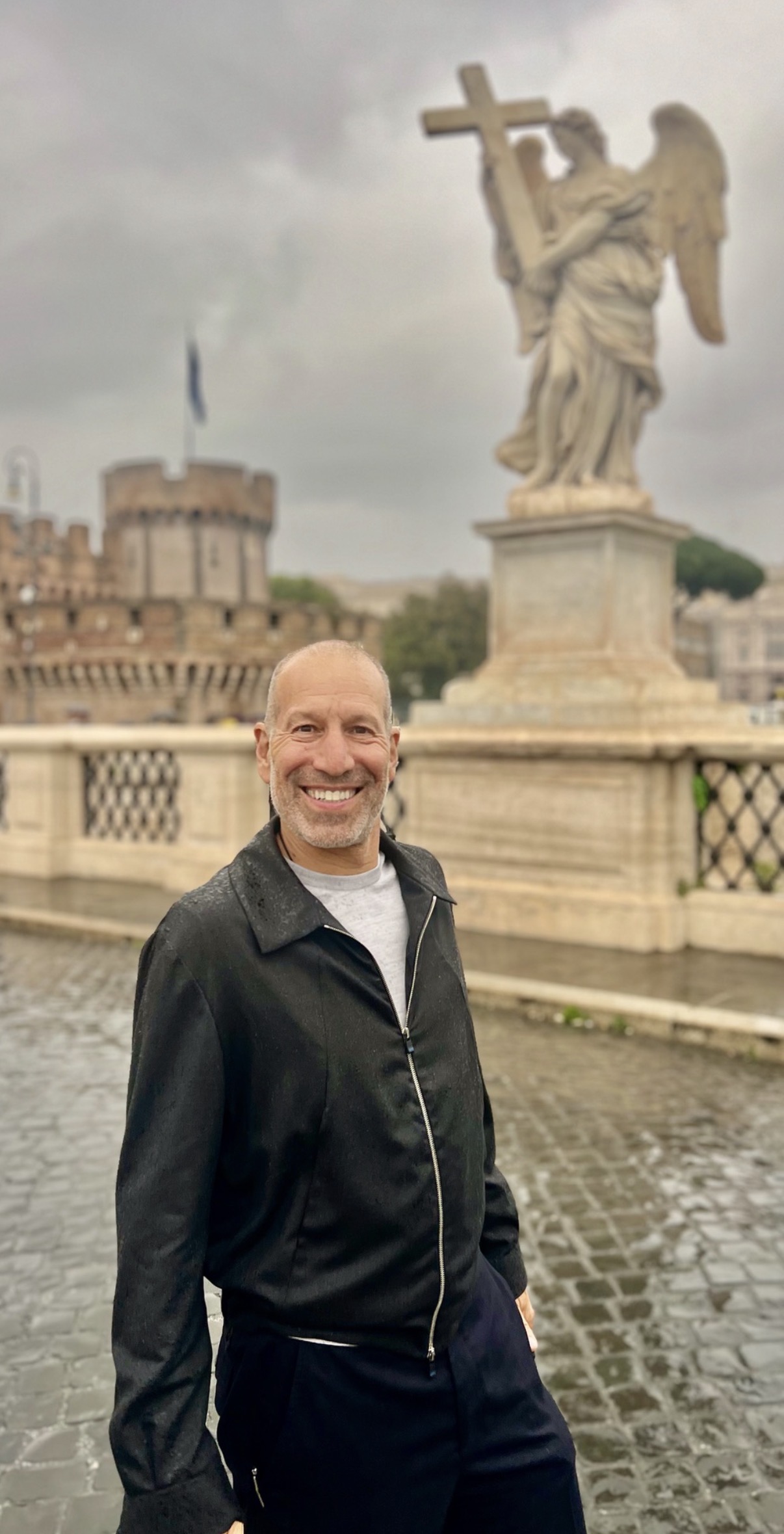 Man smiling outdoors near a fountain with a statue of an angel holding a cross, cloudy sky background.