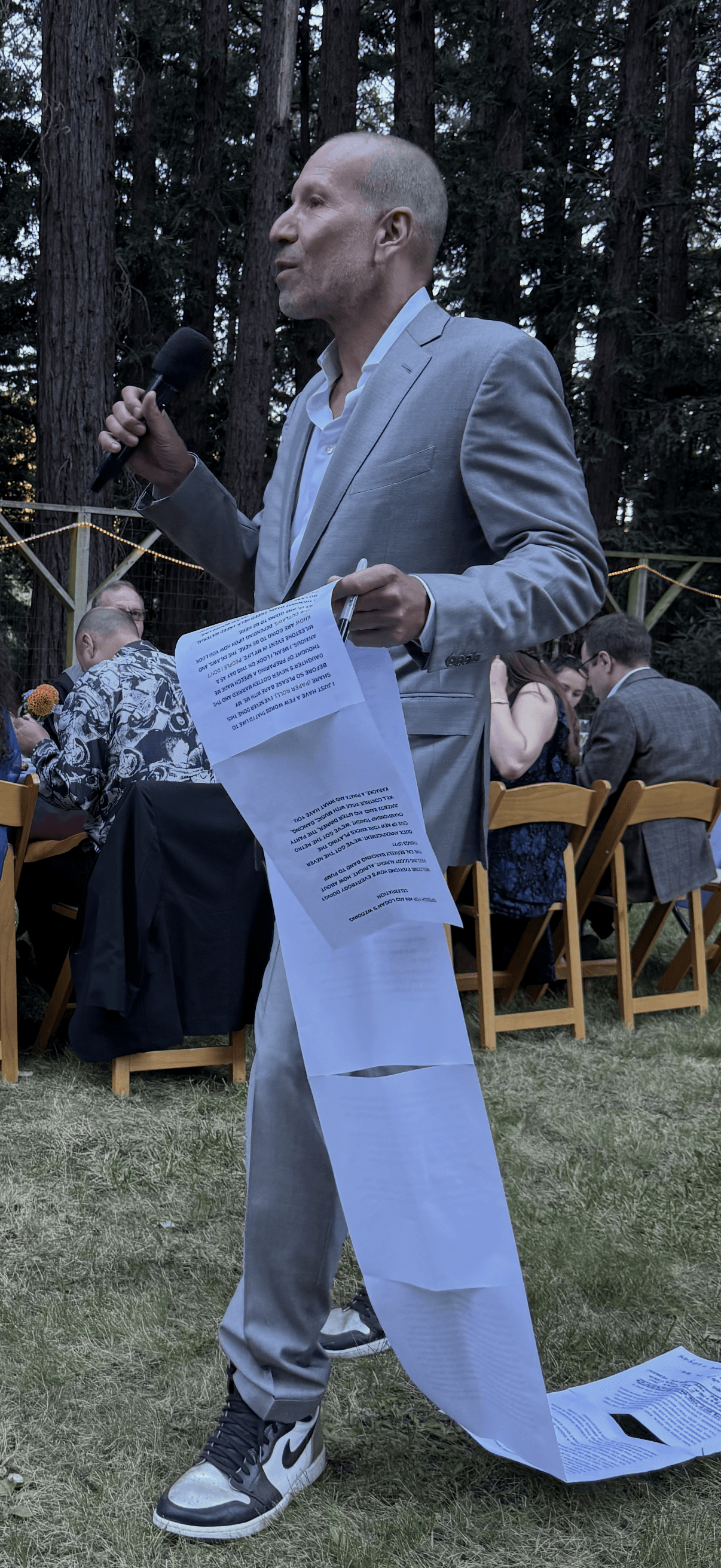Man in gray suit holding a microphone and paper, standing outdoors with seated people and trees in background.