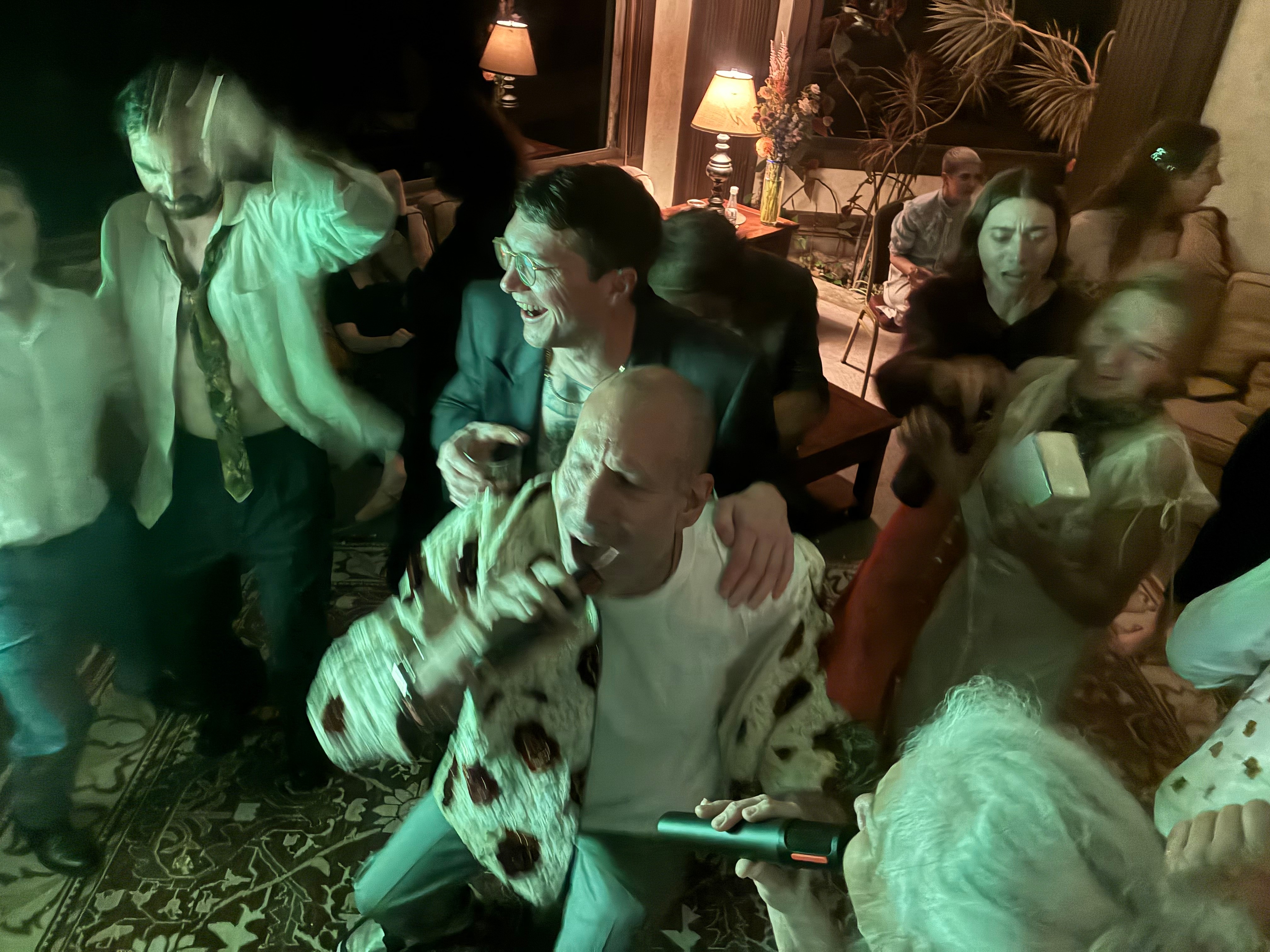 Group of people socializing indoors, some holding drinks, with warm lighting and decorative plants in the background.