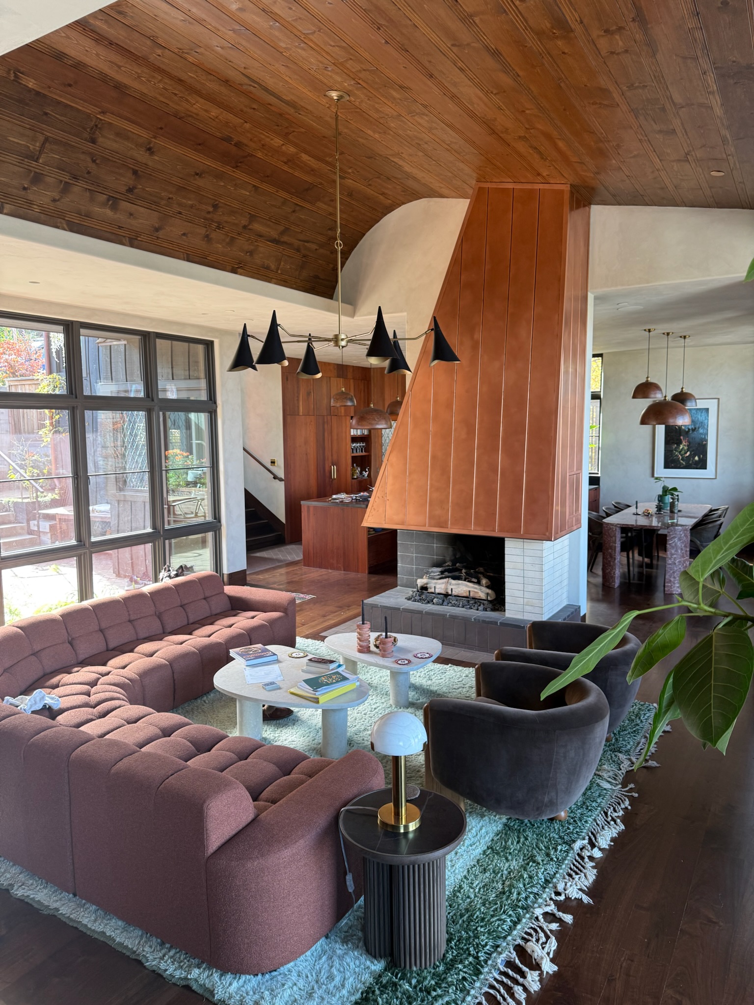 Living room with pink sectional sofa, black armchair, white coffee table, fireplace, large window, and wooden ceiling.