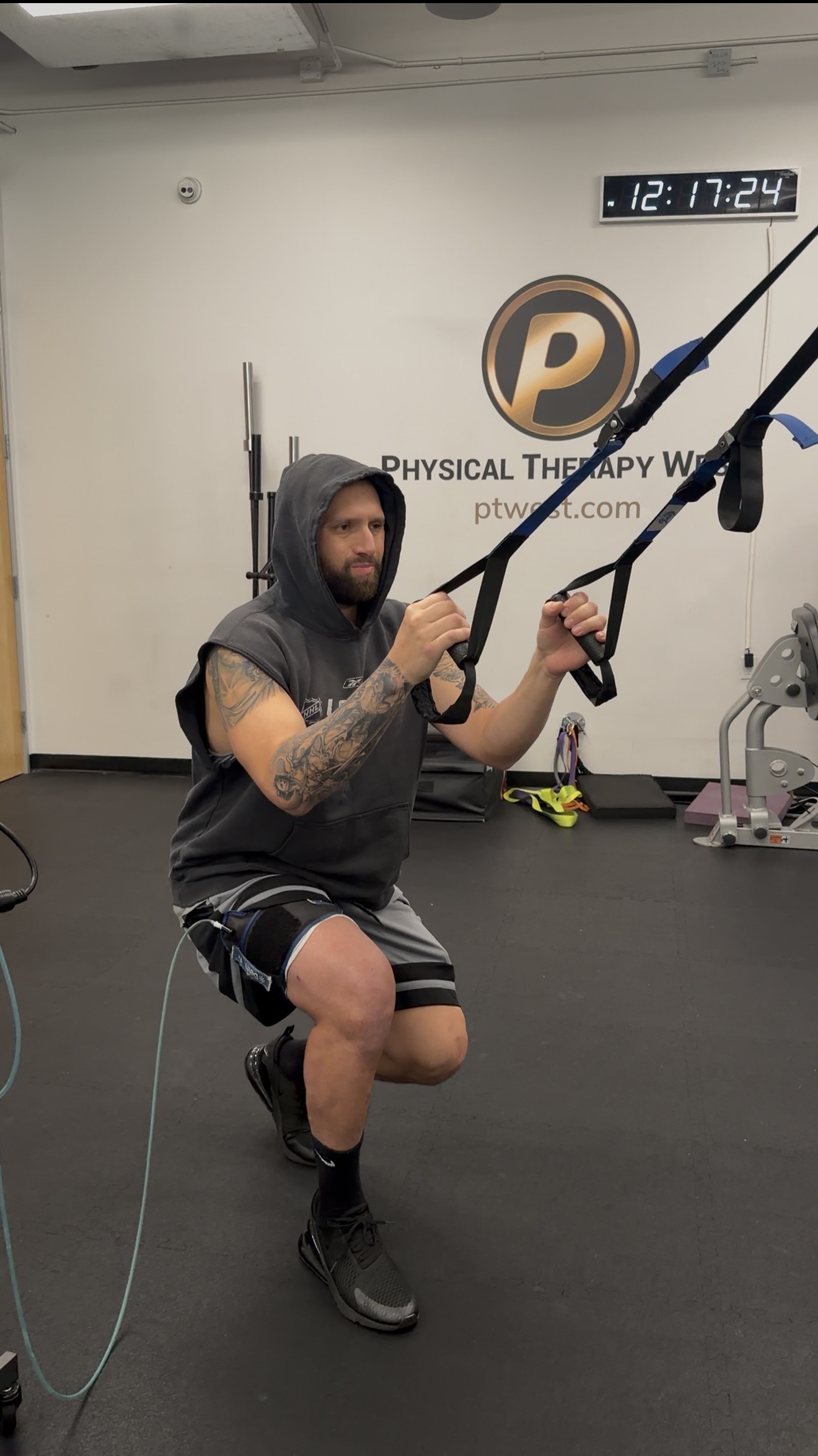 Person with tattoos in a gym holding suspension training straps, kneeling on one knee, with gym equipment in background.