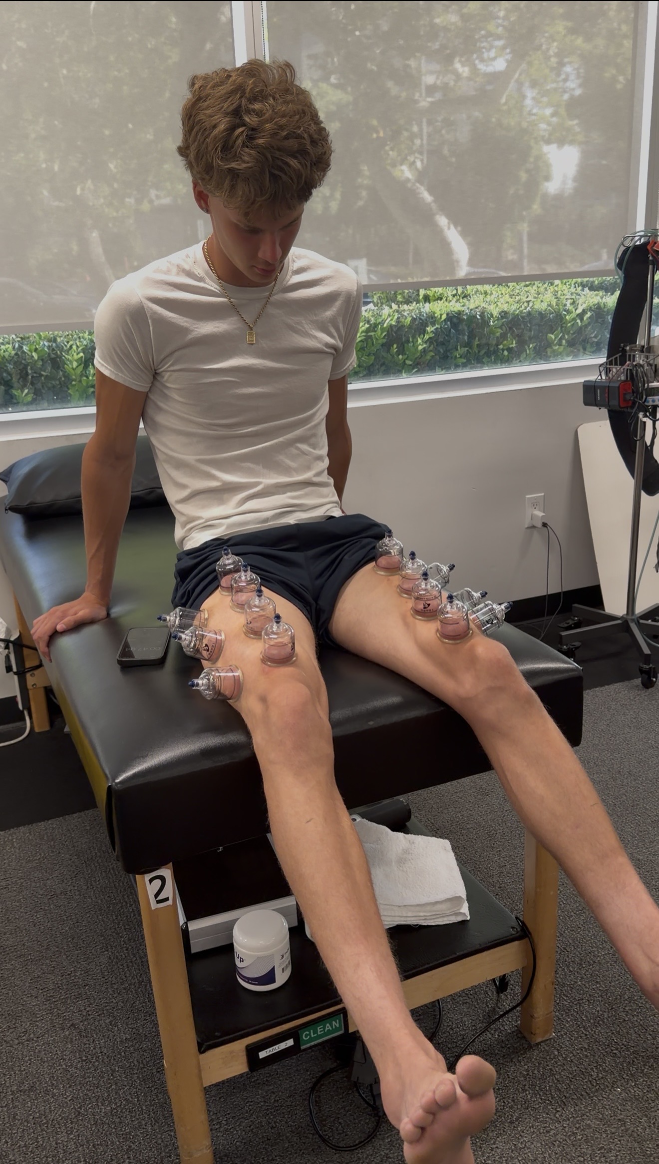 Young person sitting on a medical examination table with legs extended, covered in transparent medical dressings, in a clinical room.