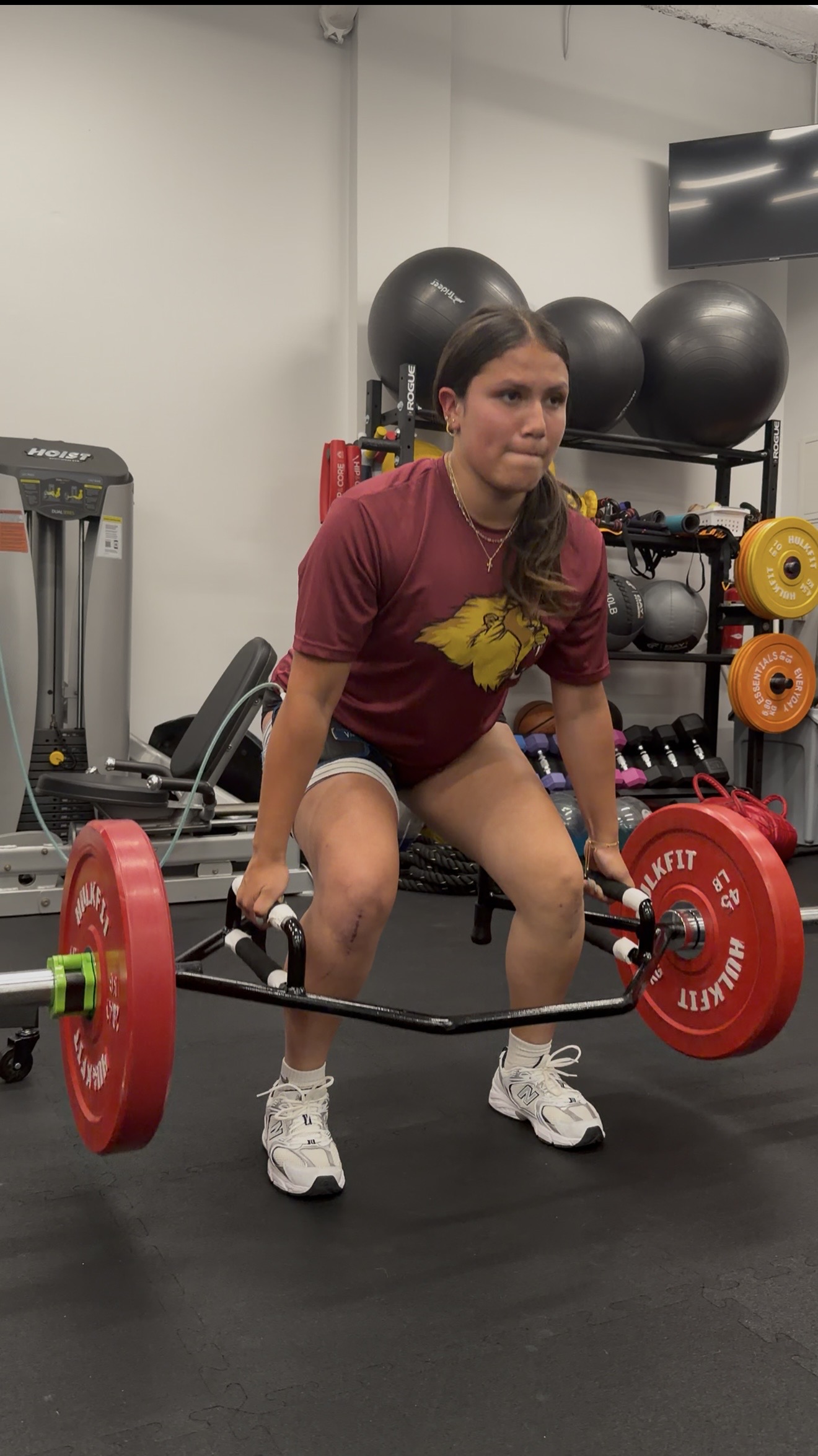 Woman in a gym lifting a barbell with red weights, surrounded by exercise equipment and black exercise balls.