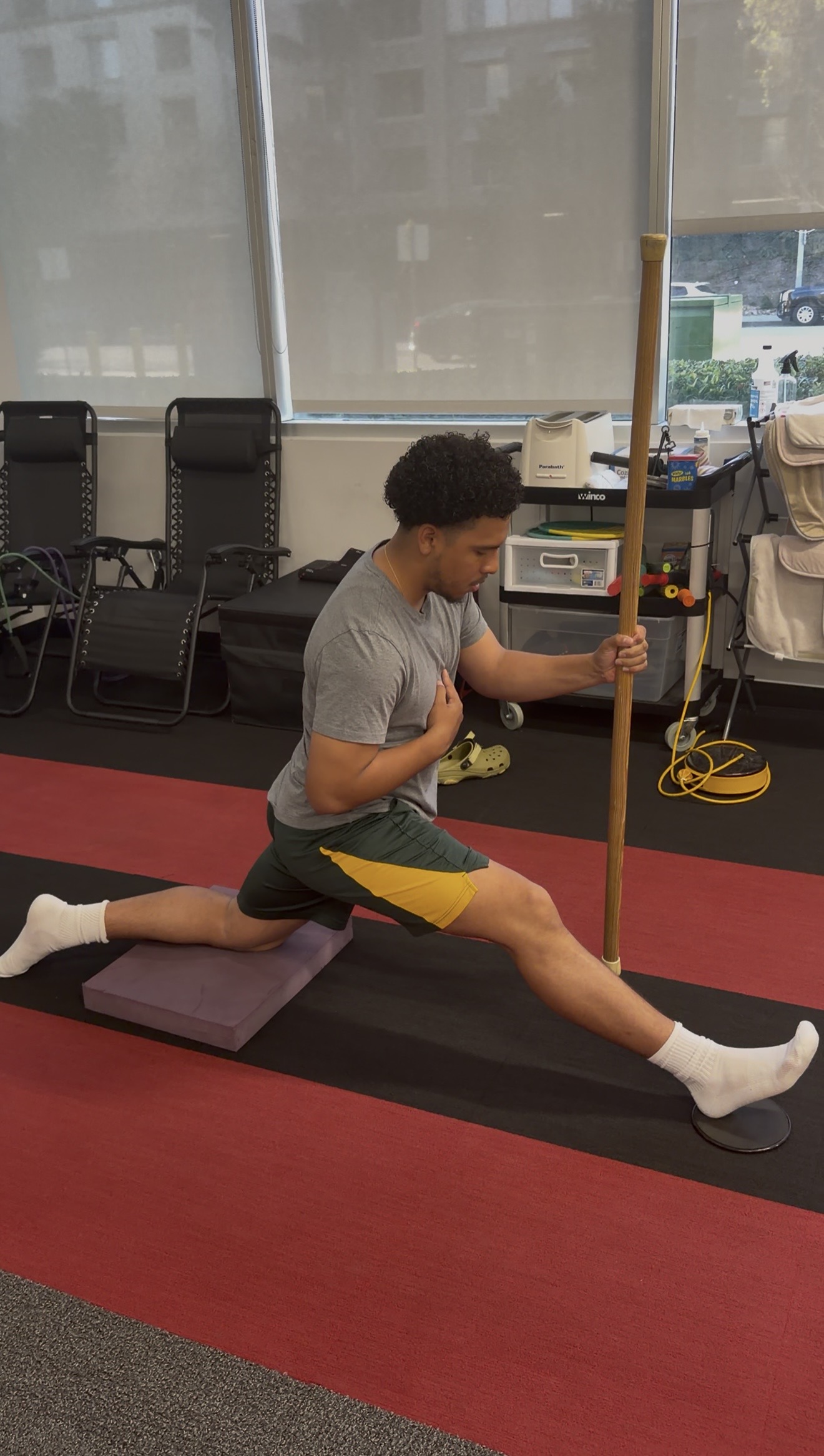 Person stretching with a pole in a gym, performing a lunge on a mat, with chairs and equipment in the background.