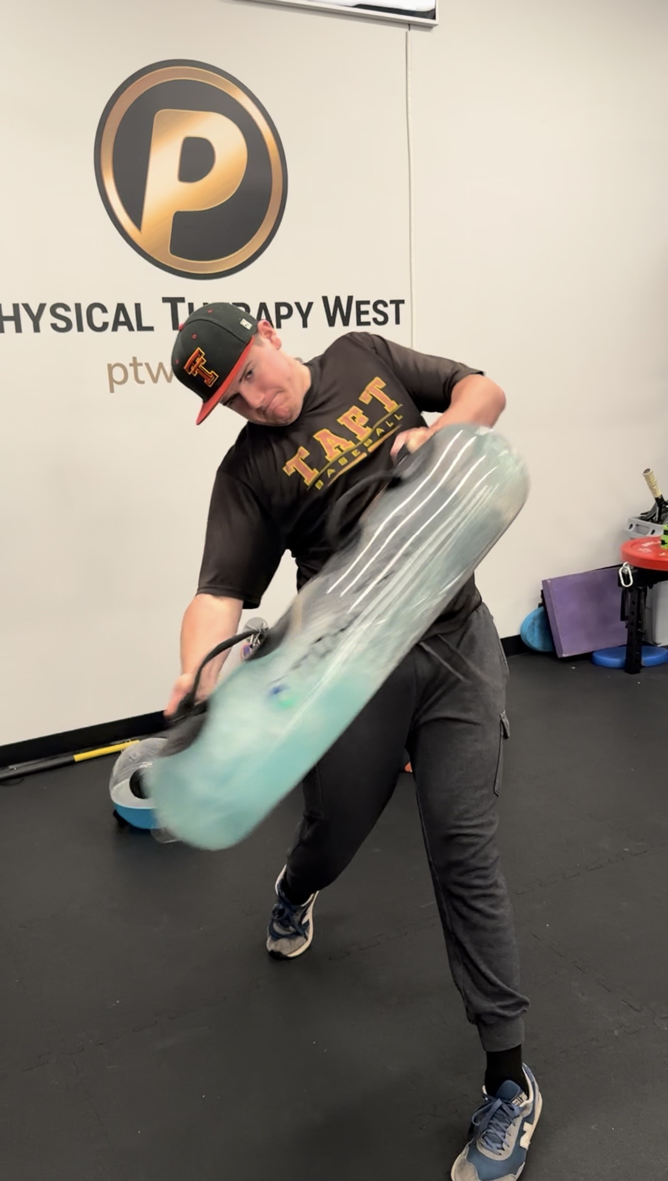 Person holding a large skateboard in a gym, with a logo and text on the wall behind them.