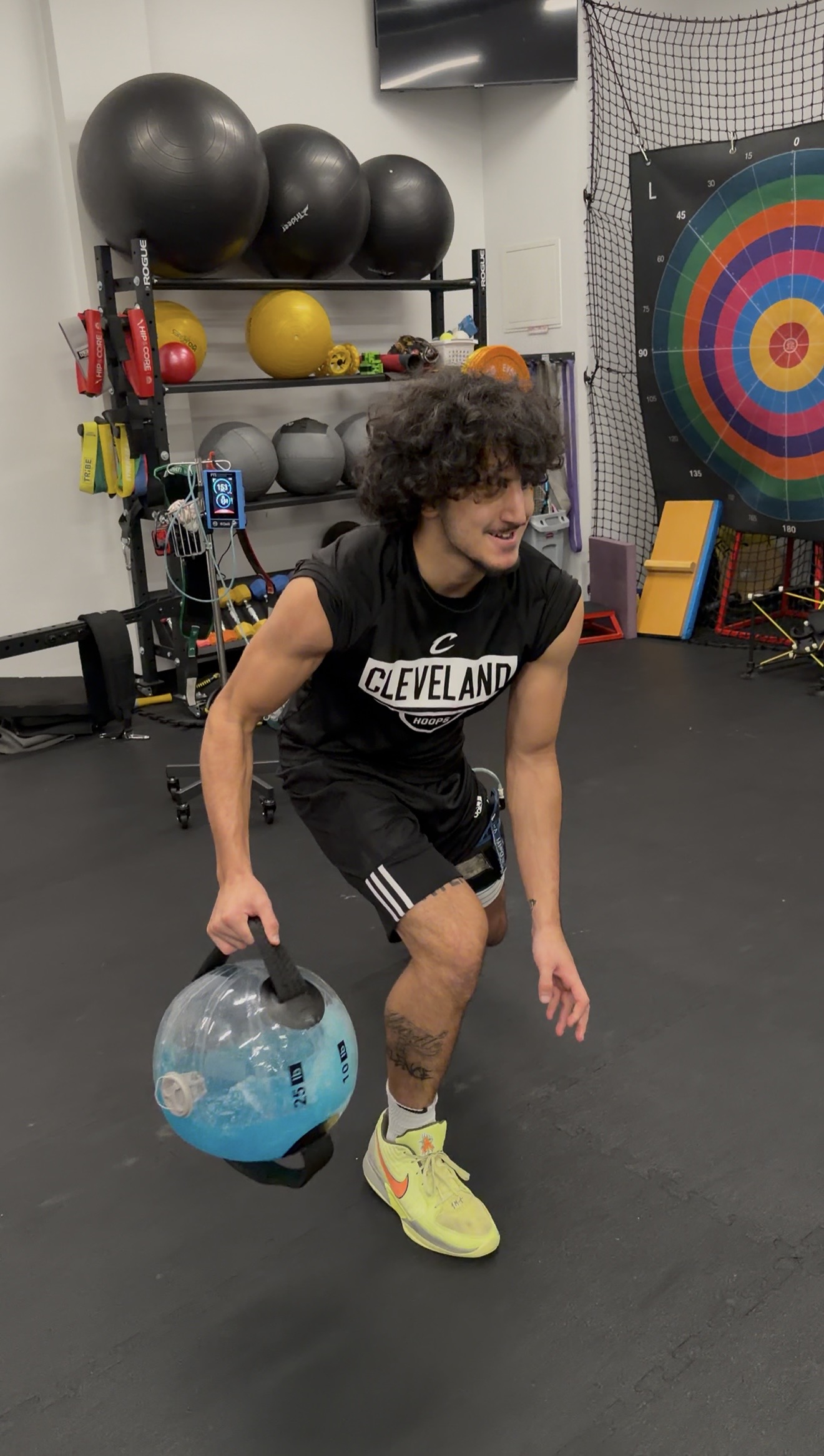 Person with curly hair in a gym holding a water jug, wearing a Cleveland shirt and yellow shoes.