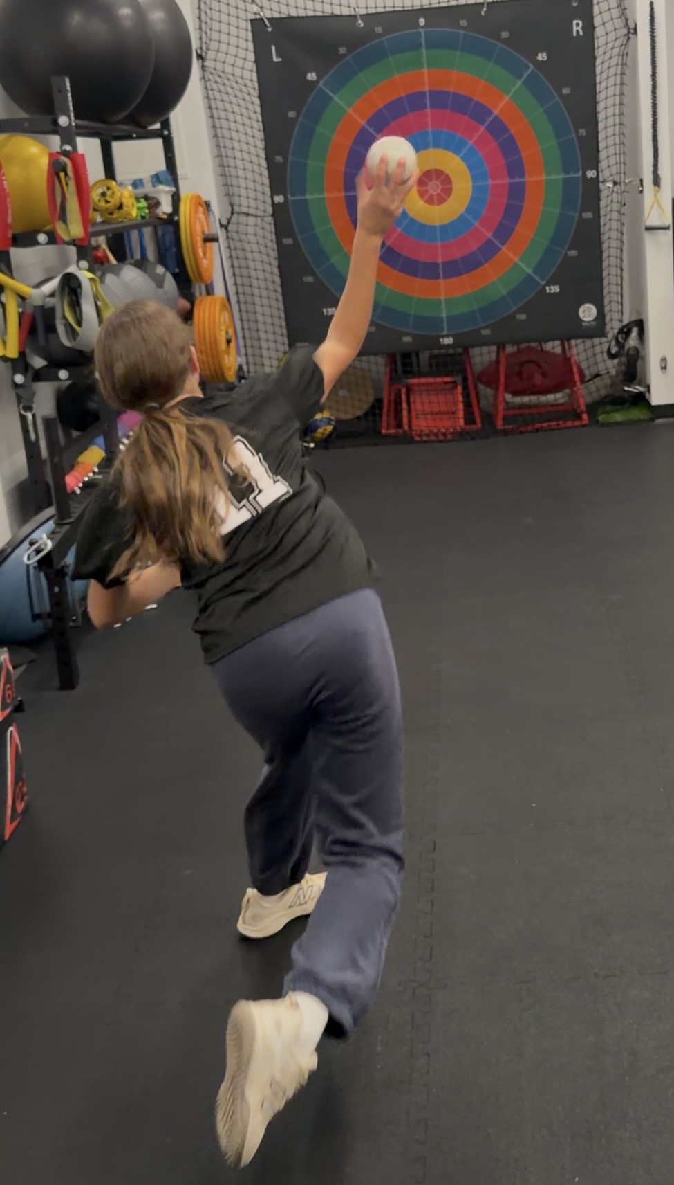 Child throwing a ball at a colorful target in a gym, with weights and equipment in the background.