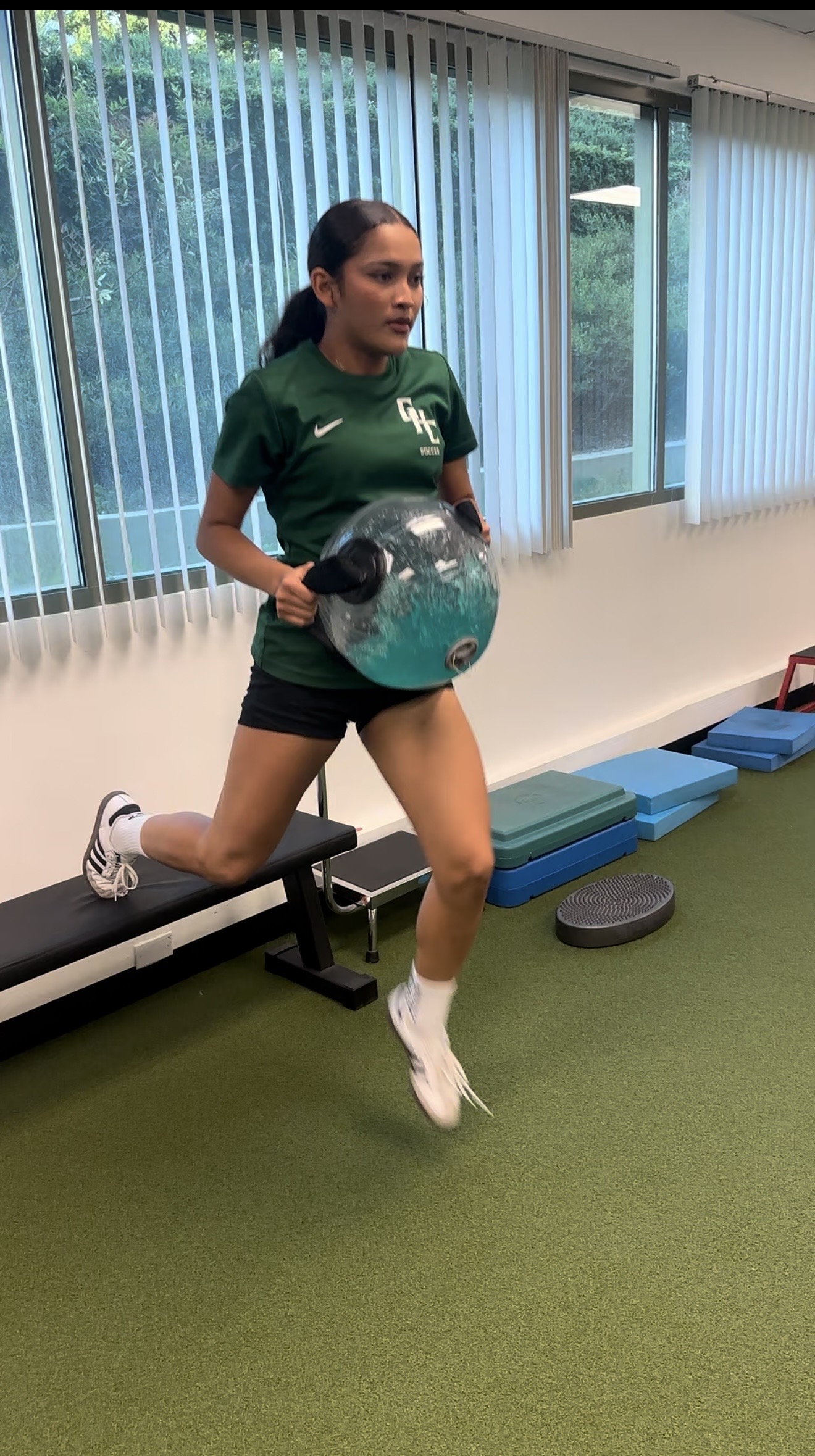 Woman in green shirt and black shorts holding a black and blue exercise ball, indoors near windows with vertical blinds.