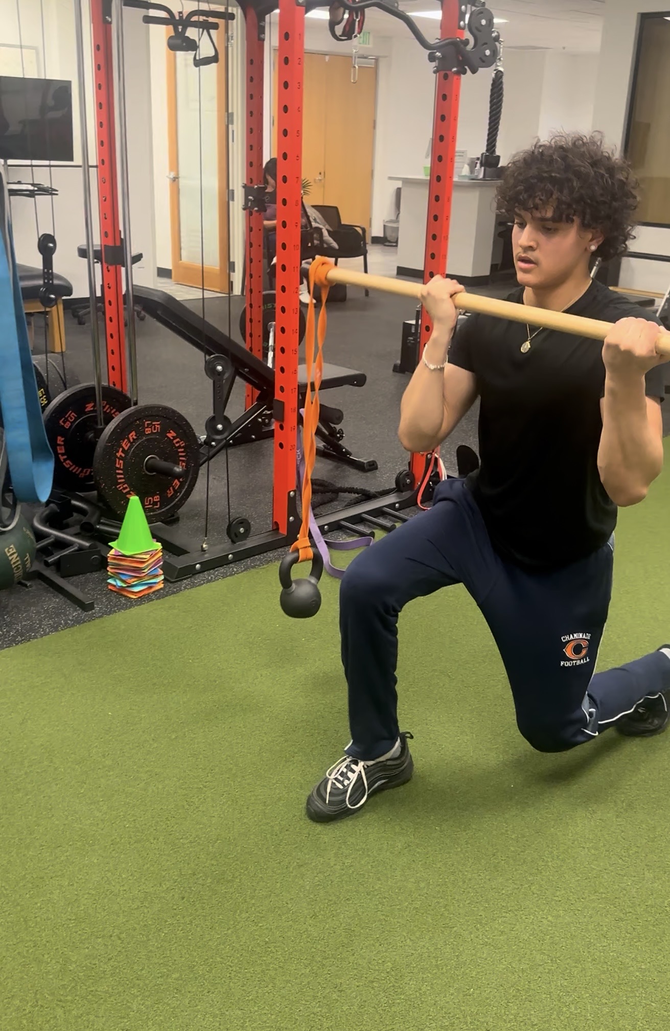 Person kneeling on green gym floor holding a wooden stick across shoulders, gym equipment in background