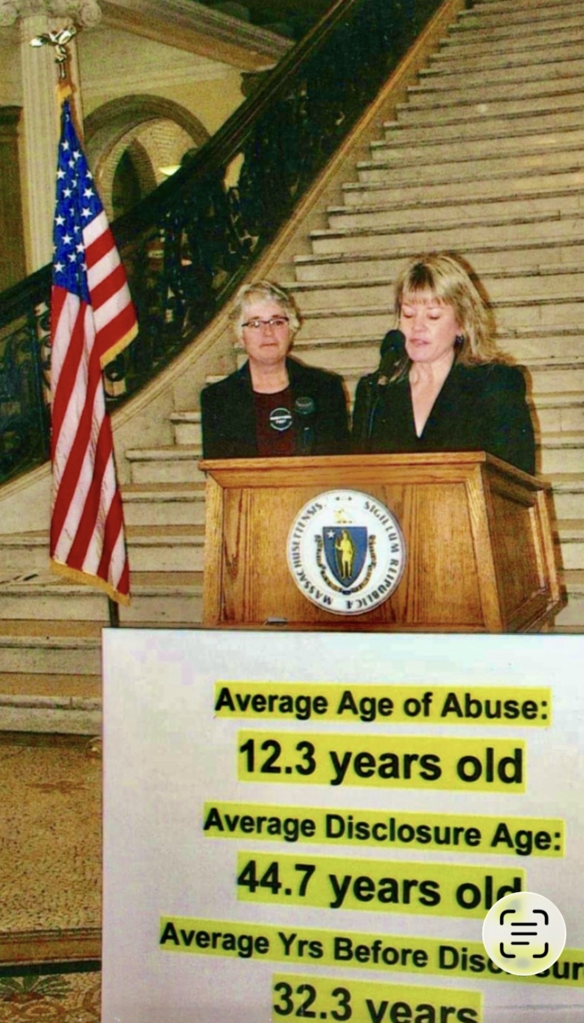 Two women stand at a podium with a seal, in front of a staircase and American flag, speaking to an audience.