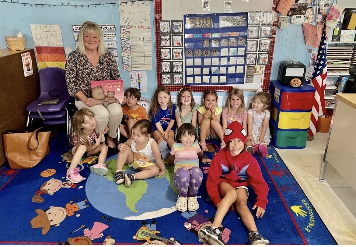 Group of children and a woman sitting on a colorful rug in a classroom, with educational materials and decorations behind them.