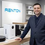 Man in a suit standing at a display counter with electronic devices in a store, Renton sign in background.